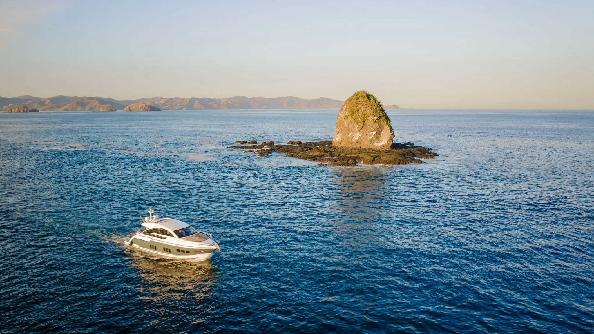 Aerial view of an Ocean Club motor yacht on flat blue water beside a rock formation on Costa Rica's Pacific coast at sunrise